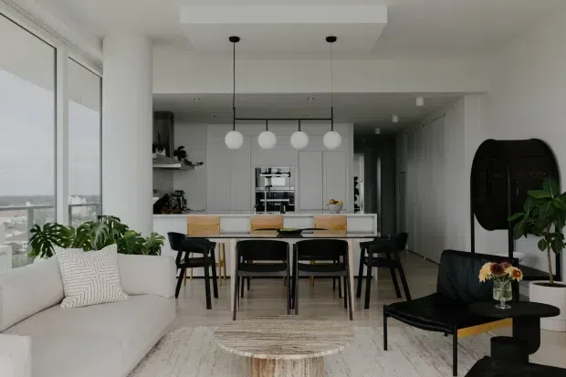A bright, open-plan apartment interior combining a living room, dining area, and contemporary kitchen. Floor-to-ceiling glazing runs along the left side, while a white kitchen with integrated cabinetry sits behind a timber dining table and black dining chairs. A white sofa, round coffee table, pendant globe lights, indoor plants, and a sculptural black lounge chair complete the refined neutral scheme.