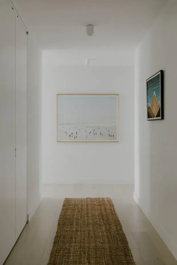 A narrow, bright white hallway or entry passage with smooth white walls and ceiling, pale timber or light neutral flooring, a long woven runner rug, minimalist flush cabinetry or doors along the left wall, recessed openings, and two framed artworks at the far and side walls. The space has a clean, restrained contemporary appearance with soft natural light.