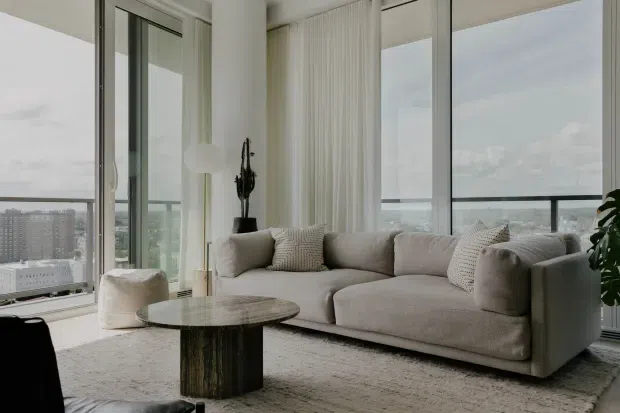 Bright contemporary high-rise living room with floor-to-ceiling glazing, expansive city and water views, sheer curtains, and a large neutral-toned sectional sofa. A round dark coffee table, textured area rug, sculptural black planter, and indoor greenery add understated contrast to the soft beige and white palette.