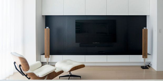 A minimalist media wall in a bright living room, featuring a wall-mounted black television set into a dark feature panel, white built-in cabinetry above and below, and matching tall timber-and-black speakers. In the foreground is a classic white leather lounge chair with a timber shell and matching ottoman. Pale timber flooring and sheer white curtains create a clean, calm interior.