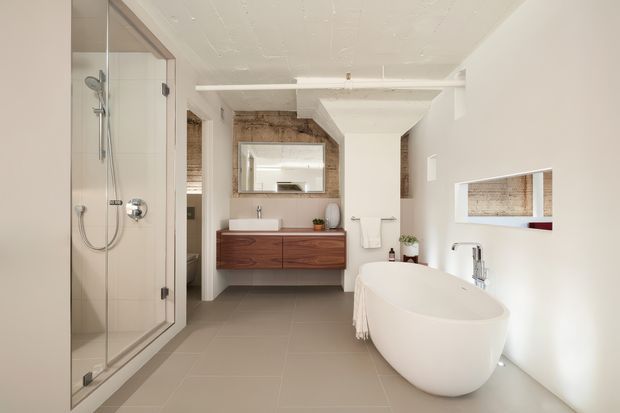 A bright contemporary bathroom with a frameless glass walk-in shower on the left, a freestanding white oval bathtub in the foreground, and a floating timber vanity with a white basin beneath a mirrored wall cabinet. Large-format grey floor tiles, white walls and ceiling, timber-look feature surfaces, chrome tapware, recessed shelving, and minimal decorative accessories create a clean, refined interior.