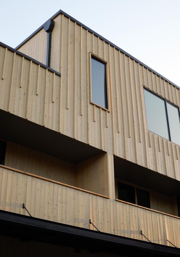 Close-up upward view of a contemporary multi-level building facade clad in pale vertical timber boards. The facade has dark contrasting trim and roof edges, projecting balcony or overhang forms, and tall rectangular windows with dark frames. The image focuses on the exterior envelope and geometric massing against a plain sky.