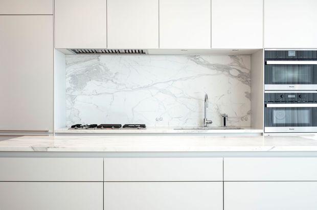 A bright, minimalist contemporary kitchen viewed straight on. Flat-panel white cabinetry spans the wall and island, with a large white marble-look veined splashback, integrated cooktop and concealed rangehood, undermount sink with a slim chrome tap, and a vertical bank of built-in stainless-steel appliances.