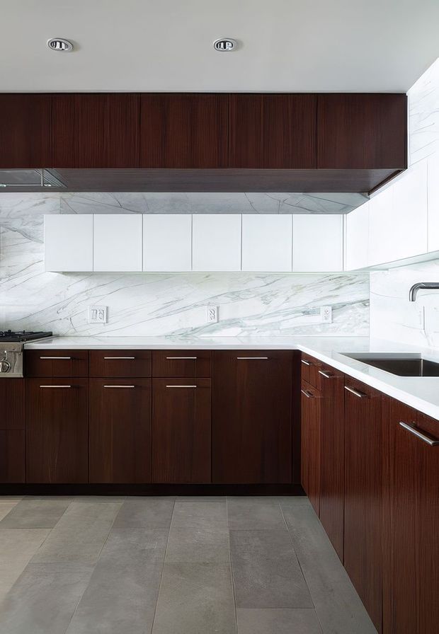 A compact contemporary kitchen with dark reddish-brown wood cabinetry, white upper cabinets, and a dramatic white marble-look backsplash and benchtop with grey veining. The kitchen includes integrated storage, a gas cooktop, undermount sink, recessed ceiling lights, and large grey stone-look floor tiles.