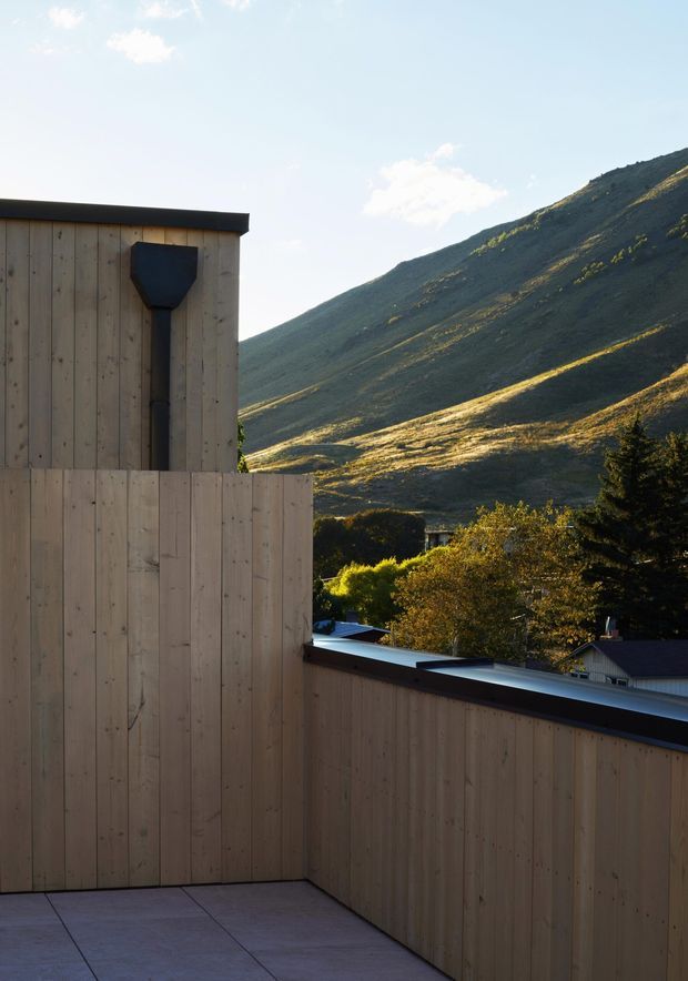 A contemporary rooftop terrace or balcony with pale vertical timber-clad walls and a dark metal coping detail. The terrace overlooks a tree-filled settlement and a steep, expansive mountain slope in warm late-afternoon light. The composition highlights the contrast between the simple architectural enclosure and the dramatic alpine landscape.