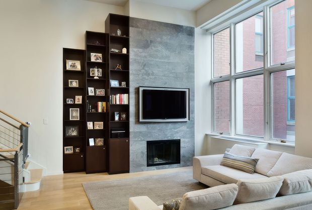 Bright contemporary living room with a large corner window, a textured grey feature wall incorporating a wall-mounted television and fireplace, and tall dark built-in shelving filled with framed photographs and books. A light sectional sofa and neutral rug sit on a warm timber floor, while a metal-and-timber stair railing is visible at the edge of the room.