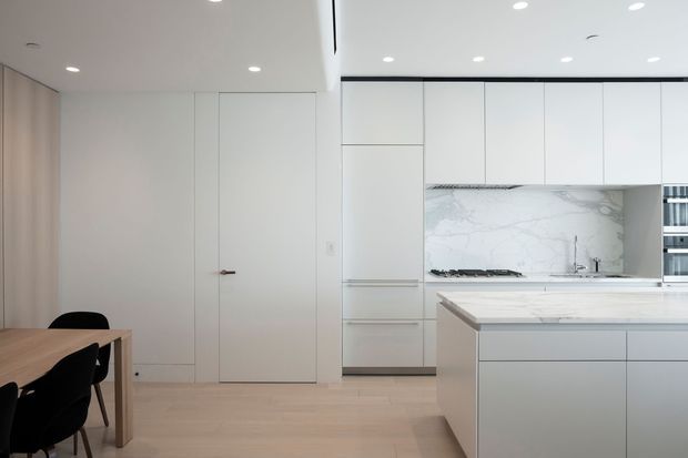 A bright, minimalist open-plan kitchen and dining area with floor-to-ceiling white cabinetry, a pale stone island and benchtop, and a marble-look splashback. A small timber dining table with dark chairs sits to the left. The room has pale timber flooring, recessed ceiling downlights, integrated appliances, and clean handleless joinery.