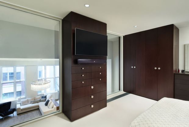 A refined modern bedroom with extensive dark timber built-in joinery. The room includes a tall dresser and wall-mounted television, full-height wardrobes with round metal pulls, pale carpet, and a partially visible bed in the foreground. A large internal glazed partition looks into a bright living area below, creating a visually connected, open interior. The palette is predominantly dark wood, white, and light neutral tones, with recessed ceiling lighting and clean-lined detailing.
