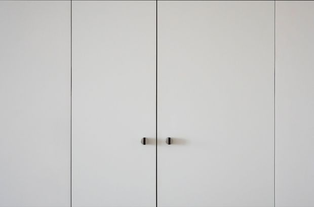 Minimal close-up of a wall of tall, flush white built-in cupboard or wardrobe doors. The doors have narrow dark seams and small dark vertical handles, creating a very clean, understated composition with no other visible room features.