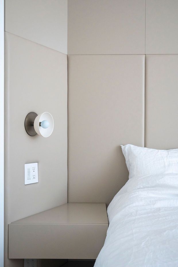 Close-up of a minimalist bedroom corner featuring a bed with white bedding, a taupe upholstered or padded wall panel system, an integrated floating bedside table, and a small wall-mounted reading light with a nearby power outlet. The composition is clean, neutral, and highly restrained.