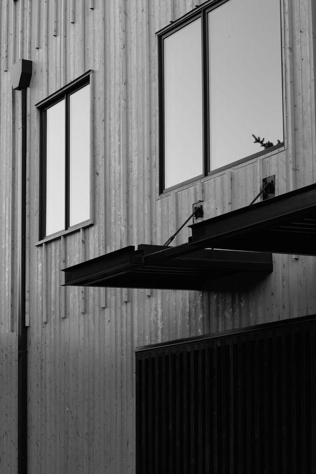 Monochrome close-up of a modern multi-storey building facade clad in vertically spaced timber boards. Tall rectangular windows have dark metal frames, and a projecting black metal awning or balcony structure creates strong horizontal and diagonal lines above a dark vertical-slat enclosure.