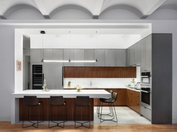 Bright contemporary kitchen with a large central island and four dark bar stools, extensive matte grey cabinetry, warm timber island and lower cabinets, a white benchtop and splashback, integrated appliances, and distinctive curved vaulted ceiling detailing. Timber flooring and a restrained grey-and-white palette create a refined, modern interior.