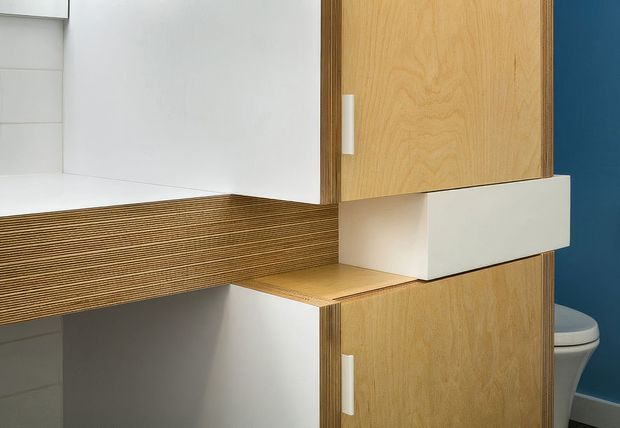 Close-up of a compact contemporary bathroom vanity and storage joinery. Light timber-faced cabinet doors and a layered timber countertop project from white cabinetry, with a white rectangular basin or storage element and a partially visible toilet against a dark blue wall. The image highlights clean geometric forms and detailed plywood edge layering.