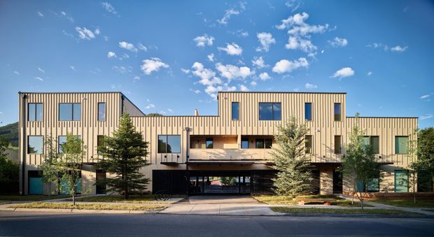 Front exterior view of a contemporary multi-storey residential building with a long, stepped facade clad in pale vertical timber-look boards. The building features varied rectangular windows, recessed balconies, a central covered carport or entry, landscaped planting, and a flat or very low-pitched roofline beneath a bright blue sky.