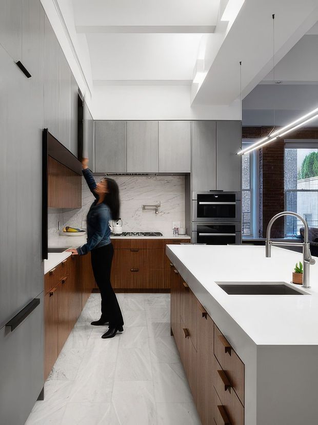 Contemporary galley-style kitchen with a large white island, integrated double sink and minimalist tapware. The room features vertical timber-grain cabinetry, tall grey cabinetry, a pale marble-look splashback and flooring, black integrated appliances, and linear pendant lighting. A glazed opening connects the kitchen to an outdoor courtyard or garden area.