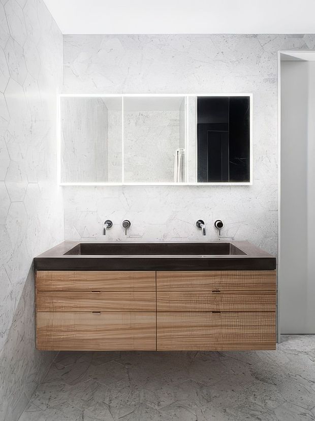 A bright, minimalist bathroom featuring a floating timber vanity with multiple drawers, a dark stone or composite benchtop with integrated double basins, wall-mounted black tapware, and a large mirrored cabinet. The walls and floor are clad in pale, veined stone-look surfaces, creating a clean contemporary setting with strong black-and-white contrast and warm timber joinery.