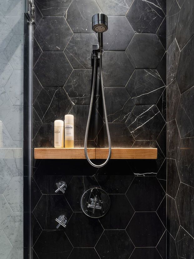 Close-up view of a compact walk-in shower clad in dark charcoal-black marble-look hexagonal wall tiles with pale veining. A brushed chrome rainfall shower and handheld shower set are mounted above a floating solid timber shelf holding toiletries, with chrome controls below. A frameless glass shower screen is visible at the left edge.