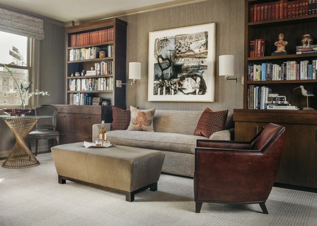A refined, traditional-style library or living room with full-height dark wood built-in bookcases, a neutral upholstered sofa, matching ottoman, and a deep brown leather armchair. Textured taupe wallcovering, framed artwork, table lamps, and a patterned area rug create a warm, sophisticated reading and conversation space.