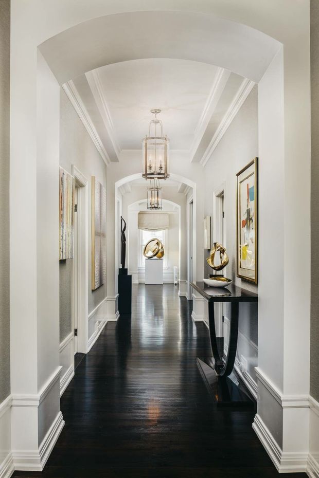 A long, elegant residential hallway viewed through a series of arched openings. The corridor has white walls, ornate layered crown moulding and baseboards, dark polished timber flooring, framed artwork, a slim black console table with sculptural gold decor, and multiple suspended lantern-style pendant lights. The bright, symmetrical composition has a refined classic character.