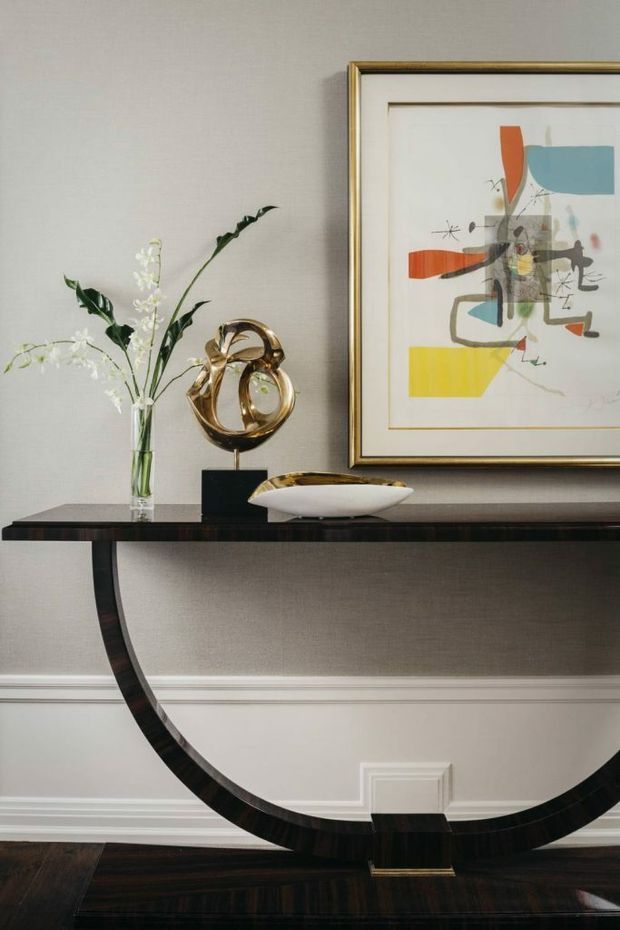 A refined interior vignette featuring a dark, glossy semicircular console table against a softly textured taupe wall with white decorative wainscoting and moulding. The console is styled with a clear glass vase of white branches, a polished gold abstract sculpture, and a shallow sculptural bowl. Above it hangs a large framed abstract artwork with a gold-toned frame and muted blue, red, yellow, grey, and black geometric forms. Dark timber flooring completes the elegant composition.