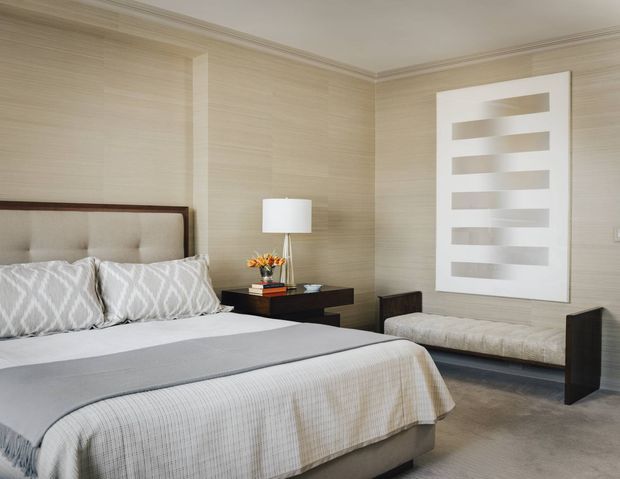A refined bedroom with a large upholstered bed, patterned pillows, a dark wood bedside table, table lamp, upholstered bench, neutral textured wallcovering, framed geometric artwork, and carpeted flooring. The palette is soft beige, cream, grey, and dark wood, creating a calm, sophisticated atmosphere.