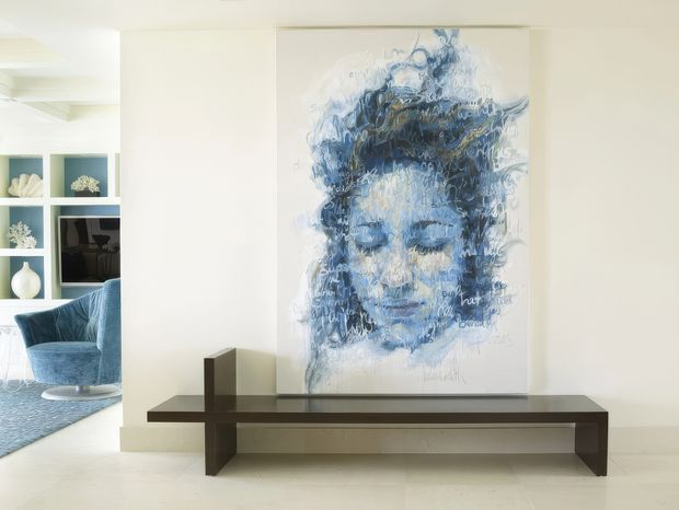 Bright contemporary interior featuring a large blue-and-white portrait artwork mounted above a long dark timber console bench. The pale walls and floor create an airy, minimalist setting, with a glimpse of an adjacent living area containing blue upholstered seating and built-in display shelving.