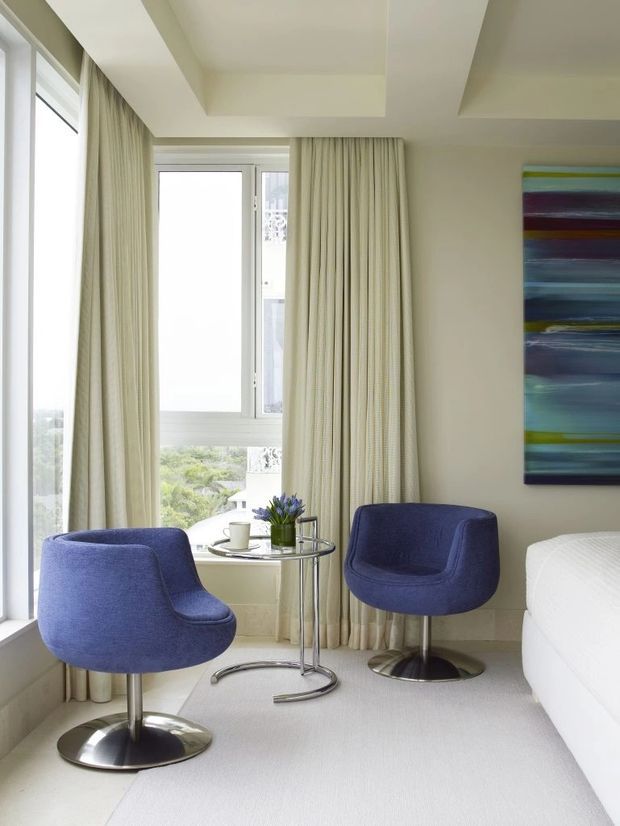 Bright, refined bedroom sitting corner with two sculptural deep-blue upholstered swivel armchairs, a small chrome-and-glass side table, and a cup of coffee beside floor-to-ceiling windows. Full-height cream curtains, pale walls, a light carpet, and a colourful abstract artwork create a calm contemporary interior.