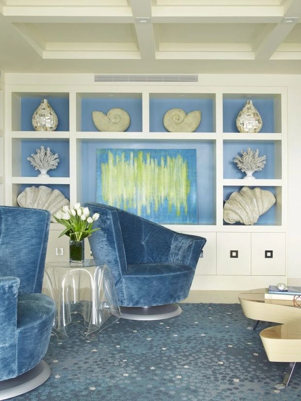 A bright, refined living room with a built-in white display and storage wall featuring blue-backed niches, shell and coral decor, and a vivid abstract artwork. Blue velvet swivel armchairs, a transparent acrylic side table, a patterned blue rug, and a sculptural light coffee table create a coastal-inspired, polished interior. The ceiling has white exposed beams and recessed coffers.