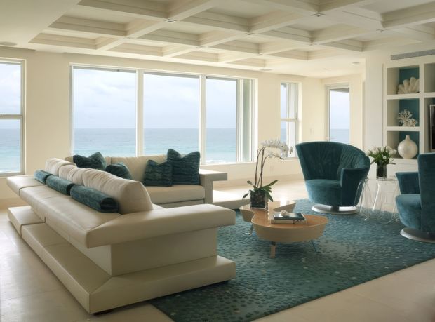 Bright, spacious coastal living room with expansive ocean-facing windows, a white sectional sofa, teal upholstered armchairs, a round timber coffee table, and a patterned teal rug. The room features a distinctive coffered ceiling, pale flooring, built-in shelving, and an airy refined interior overlooking the sea.