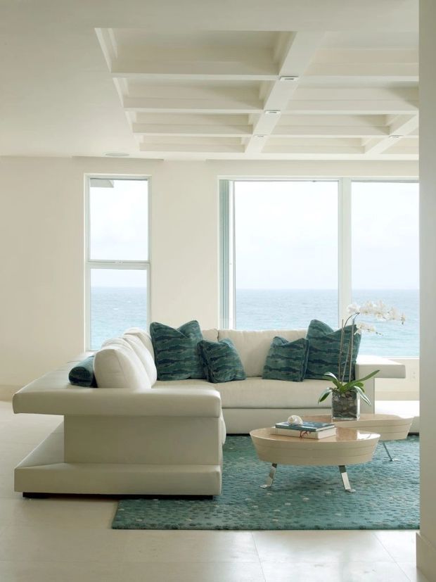 Bright coastal living room with a large cream sectional sofa positioned in front of expansive ocean-facing windows. The room features a sculptural white coffered ceiling, pale walls, a textured teal area rug, coordinating teal cushions, and a light wood oval coffee table with an orchid arrangement. The space has a clean, airy, refined aesthetic and clear ocean views.