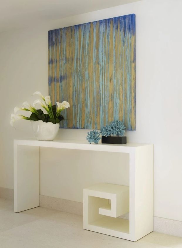 A minimalist interior vignette featuring a white console table against a plain white wall, with a small geometric white stool or side table tucked beneath it. A large abstract artwork in blue, teal, and ochre hangs above, complemented by white flowers in a sculptural vase and blue-toned decorative objects. The space has a pale stone or tile floor and a calm, neutral palette with vivid artwork accents.