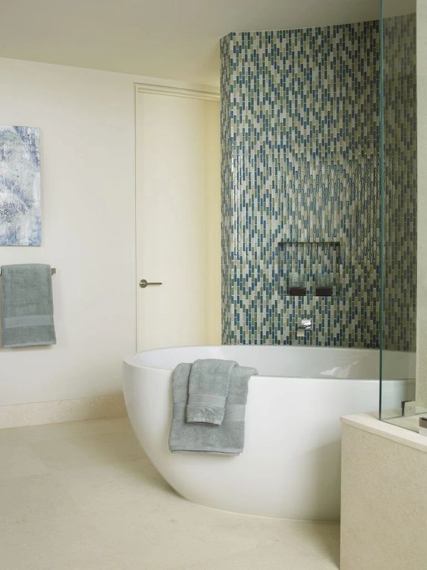 Bright contemporary bathroom featuring a freestanding white bathtub, a full-height glass shower screen, and a striking mosaic-tile feature wall in muted blue, green, grey, and cream tones. The room has pale neutral walls and floor finishes, minimal white cabinetry or vanity elements, brushed metal tapware, and soft grey towels. A closed door and wall-mounted towel rail are visible, creating a clean, refined spa-like atmosphere.
