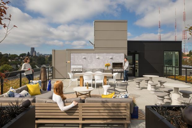 A spacious rooftop terrace designed for entertaining, with outdoor lounge seating, dining furniture, sculptural stools, large planters, and a built-in outdoor kitchen or bar area. The terrace has contemporary dark cladding, a tiled feature wall, metal balustrades, and broad city views beneath a partly cloudy sky.