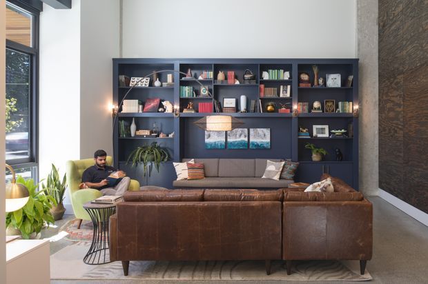 A stylish, high-ceiling living room centred on a large built-in dark shelving and display wall. The room features a substantial brown leather sectional sofa, grey upholstered seating, a green lounge chair, patterned cushions, indoor plants, framed artwork, and warm pendant lighting. Tall black-framed windows bring in abundant natural light, while a textured dark feature wall adds contrast. The overall design is contemporary with industrial influences and a warm, layered atmosphere.