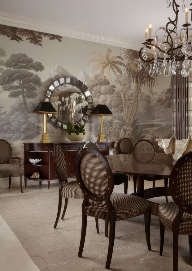 Elegant formal dining room with a dark wood dining table and cane-backed dining chairs, set against a large monochromatic tropical landscape mural. A crystal chandelier, matching table lamps, a mirrored sideboard, ornate mirror, orchids, and sculptural shell decor create a richly layered, refined interior. Pale stone-patterned flooring and neutral walls balance the dark furniture and dramatic wallpaper.