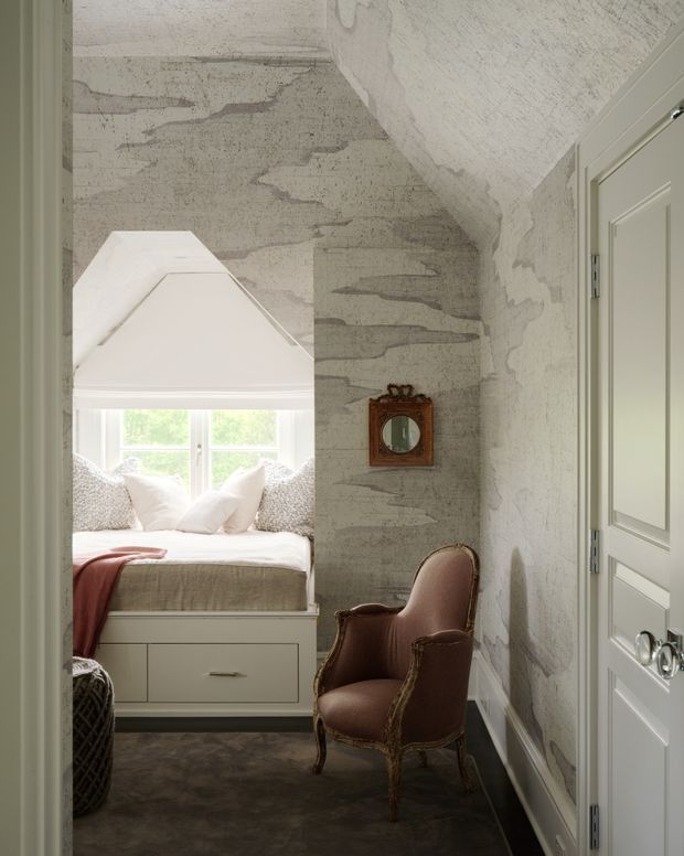 A narrow, softly lit bedroom nook or reading area with a built-in window seat, white cabinetry, layered cushions, and a rust-coloured throw. The space features heavily textured grey plaster walls and ceiling, a vintage upholstered armchair, a small ornate wall mirror, paneled white doors, and dark flooring. The overall feel is intimate, historic, and richly textured.