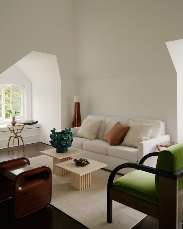 A refined contemporary living room with a white upholstered sofa, sculptural green accent chairs, dark polished wood lounge chairs, and a light timber coffee table arranged on a large neutral rug. The room has warm greige walls, white built-in cabinetry beneath a multi-pane window, restrained decorative objects, and an angled architectural ceiling detail. Natural daylight creates a calm, sophisticated atmosphere.
