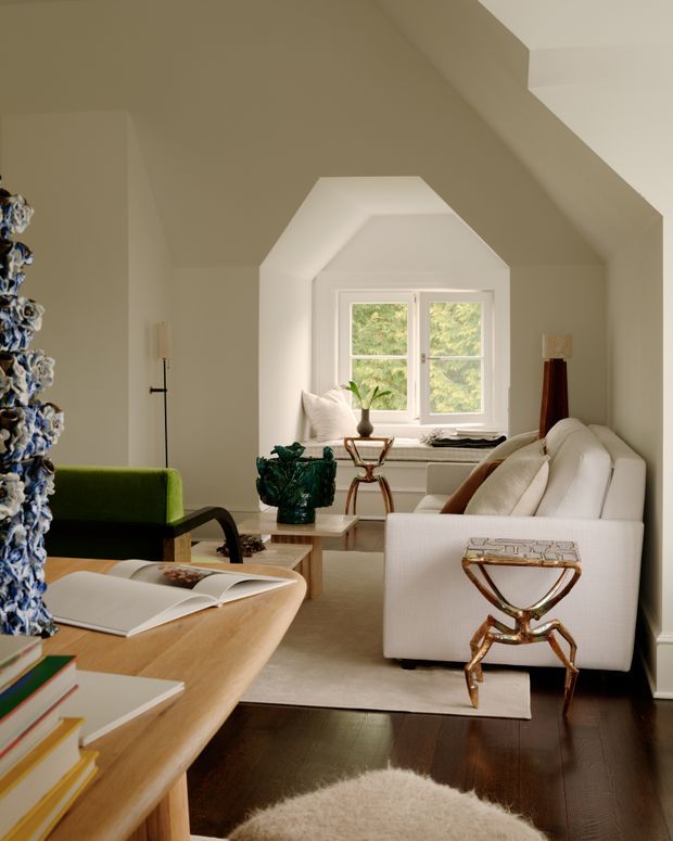 A refined living room with a white upholstered sofa, green accent chair, sculptural metallic side tables, and a pale wood coffee table and desk-like surface. The room features dark timber flooring, a large pale area rug, cream walls, and a distinctive recessed window seat beneath a faceted, angled ceiling alcove. Natural light enters through the double window, while decorative objects and books add colour and texture.