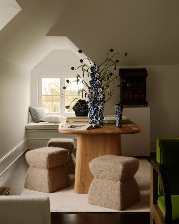 A warmly styled dining area in a compact room with a sculptural timber pedestal dining table, upholstered boucle dining stools, and a green upholstered chair. A large floral arrangement, books, and decorative vessels sit on the table. The room features white walls, a deep angled ceiling, a window seat with cushions, timber flooring, and a dark decorative cabinet against the rear wall.