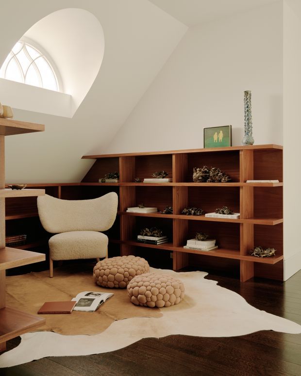 Warm, refined study or library corner beneath a sculptural sloped ceiling. A large built-in dark timber shelving unit displays books, ceramics and decorative objects, with a compact desk and sculptural boucle lounge chair nearby. A high arched window brings in soft natural light, while a sculptural pendant, organic pebble-shaped ottomans and an irregular hide rug create a sophisticated, tactile interior.