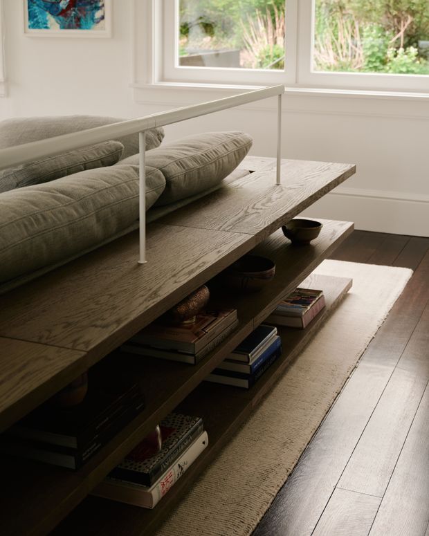 A warmly lit living room vignette featuring a long, low, dark timber open-shelf coffee table filled with books and decorative bowls. A sculptural grey sofa sits behind it, with a slim linear light or raised rail spanning the table. Large white-trimmed windows overlook leafy greenery, while a neutral rug rests over dark timber flooring. The interior has a refined, understated contemporary character with natural textures and abundant daylight.