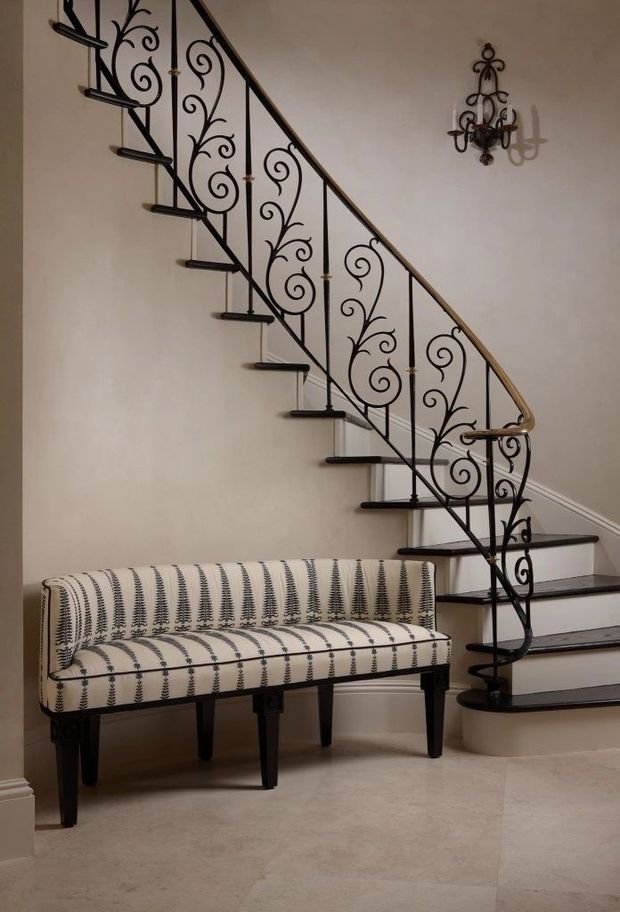 Elegant interior stairway with a sweeping curved staircase, dark timber treads, and ornate black wrought-iron balustrade featuring scrolling botanical motifs. A patterned upholstered bench sits beneath the stairs against warm off-white walls, with a decorative wall sconce and pale stone or tile flooring.