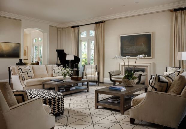 Elegant, generously proportioned living room with a refined neutral scheme. The room features several upholstered armchairs and sofas, two dark wood coffee tables, a geometric patterned area rug, a black grand piano, tall curtained windows, a fireplace mantel with decorative objects, wall panelling and framed artwork. The overall look is polished and classic with contemporary geometric accents.