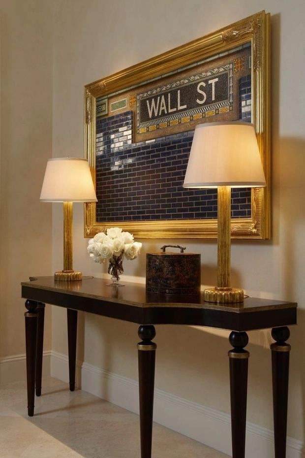 A refined entryway vignette featuring a dark wood console table with turned legs, two brass table lamps, a floral arrangement, and a decorative vessel. Above the console is a large ornate gilded frame containing a blue tile mosaic with the text “WALL ST.” The space has warm neutral walls, white skirting, and pale stone or tile flooring.