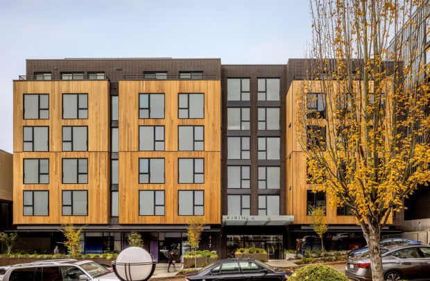 Street-level view of a contemporary multi-storey apartment or mixed-use building. The facade combines warm vertical timber-look cladding, dark brown brickwork, and extensive black-framed rectangular windows, with ground-floor commercial glazing and landscaped street planting in front. Parked cars and autumn trees occupy the foreground.