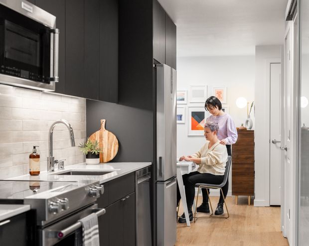A contemporary galley kitchen viewed toward an adjoining dining and hallway area. The kitchen features matte dark cabinetry, stainless-steel appliances, a light tiled splashback, pale stone benchtops, and timber flooring. Two people are seated and standing near a small dining or work table in the background, with framed artwork and timber furniture beyond.