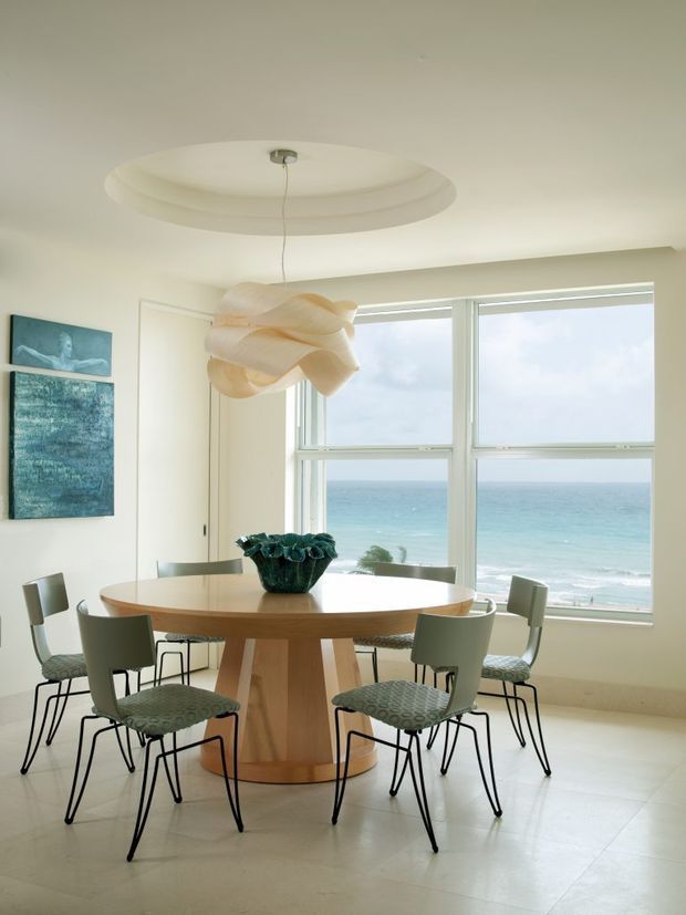 Bright coastal dining room with a round light-wood dining table and six patterned upholstered chairs, positioned in front of large windows overlooking the ocean. The room has pale walls, a light tiled floor, a sculptural cream pendant light, a recessed circular ceiling detail, and abstract wall art.