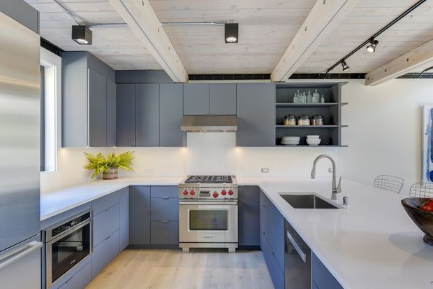 Bright contemporary galley-style kitchen with matte blue-grey cabinetry, white benchtops and splashback, stainless-steel appliances, an island with integrated sink, and exposed pale timber ceiling beams. Open shelving displays crockery and jars, while black track lighting and recessed fixtures add contrast.