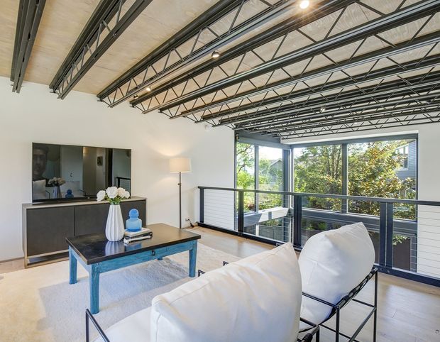 Bright contemporary living room with white walls, pale timber flooring, expansive black-framed glazing, and a dramatic exposed steel roof structure with open trusses. A white sofa and lounge chairs face a dark coffee table, while a black built-in media cabinet and large artwork occupy the rear wall. The elevated-looking glazing opens toward leafy greenery and a neighbouring modern facade.