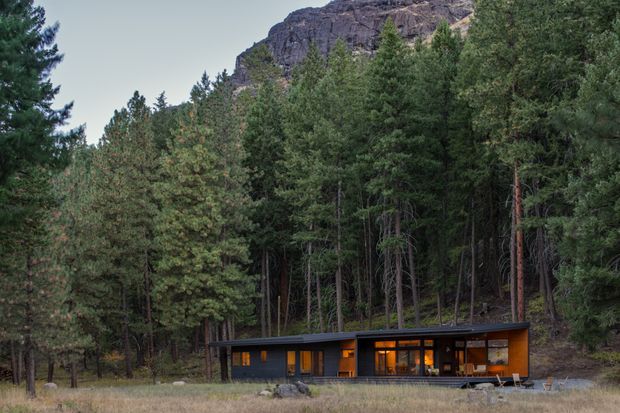 A low, single-storey modern cabin set at the edge of a dense pine forest, with a dark horizontal roofline, warm timber-toned illuminated interiors, extensive glazing, and a rugged rocky mountain outcrop rising behind the trees.